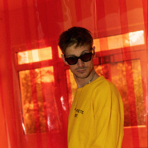 Halbnahe Seitenansicht eines Mannes vor rot-orangem Hintergrund mit transparenten PVC-Streifen. Er trägt ein leuchtend gelbes Longsleeve mit schwarzem Textprint und eine schwarze Sonnenbrille. Das Licht taucht die Szene in warmes, kontrastreiches Farbspiel.
