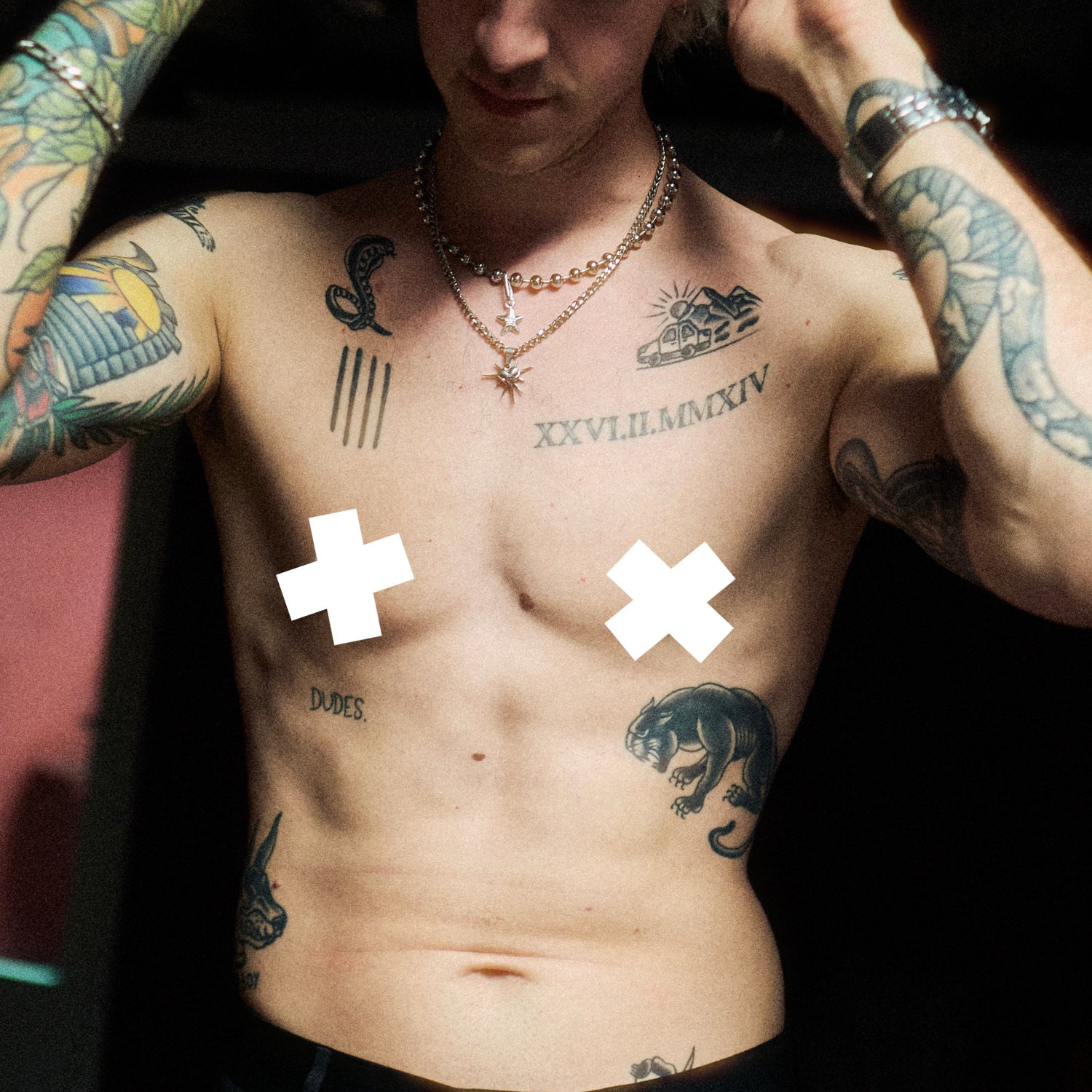 Nahaufnahme eines tätowierten Oberkörpers mit verdeckten Brustwarzen durch weiße Symbole: Sichtbar sind verschiedene Tattoos, u. a. ein „dudes.“-Schriftzug auf den Rippen, ein Panther auf der rechten Körperseite, eine römische Zahl auf der Brust sowie eine stilisierte Sonne und ein Autodach. Arme und Hals mit auffälligem Schmuck und weiteren Tattoos.