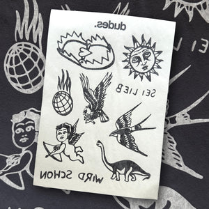 Nahaufnahme eines Tattoo-Stickerbogens auf schwarzem Stoff mit weißen Illustrationen. Der Bogen zeigt Motive im Flash-Tattoo-Stil: zwei Vögel, eine Sonne mit Gesicht, ein Herz mit Flamme, ein Dino, ein flammender Globus, ein fliegender Engel mit Pfeil und Bogen sowie die gespiegelten Schriftzüge „SEI LIEB“, „WIRD SCHÖN“ und „dudes.“.